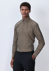Camicia a maniche lunghe in motivo marrone con colletto strutturato, caratterizzata da disegni geometrici in un colore più chiaro, abbinata a pantaloni neri.