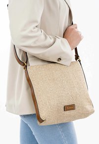 Borsa a tracolla beige intrecciata con superficie texturizzata, dettagli in pelle marrone e tracolla regolabile. Una placca con il logo è visibile sulla parte frontale.