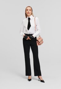 White button-up shirt with a black tie, black tailored pants, black gloves, and a pink handbag with chain accents. Black high-heeled shoes.