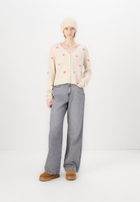 Cárdigan beige con patrones de corazones rosas, escote en V y botones frontales, combinado con unos pantalones de mezclilla de pierna ancha gris de talle alto y zapatos de plataforma beige.