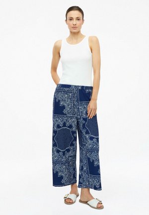 Vrouw staand, gekleed in een witte mouwloze top, blauwe wijde broek met witte paisley-patronen en witte slidesandalen met gespen.