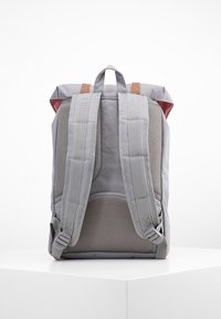 Harmaa reppu, jossa on pehmustettu verkkomateriaali selkäpuolella ja hihnoissa. Sisältää ruskeita nahkaosia ja punaisen vuorauksen. Yksinkertainen, suorakulmainen muotoilu.