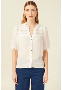 Blusa bianca a maniche corte con chiusura a bottoni sul davanti, con dettagli in pizzo, colletto e tessuto strutturato.