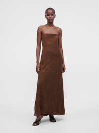 Femme portant une robe longue marron sans bretelles avec une broderie florale subtile, debout en sandales noires à talons sur un fond clair uni.