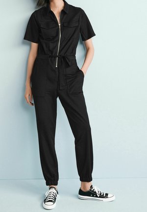 Czarny kombinezon na zamek błyskawiczny z krótkimi rękawami, dwoma przednimi kieszeniami, regulowanym paskiem w talii i zwężanymi nogawkami, połączony z czarno-białymi trampkami.