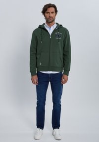 Sudadera con capucha y cremallera verde, con bolsillos delanteros y logo bordado. Combinada con vaqueros azules y zapatillas blancas. Textura suave.