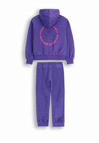 Fioletowa bluza z kapturem, z różowym okrągłym wzorem tekstowym na plecach; dopasowane fioletowe spodnie dresowe z ściągaczami na nogawkach.
