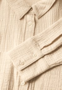 Světle béžová bavlněná košile s texturovaným povrchem, s špičatým límcem a manžetami na knoflíky. Knoflíky jsou kulaté a bílé.