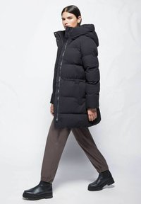 Schwarze gepolsterte Jacke mit Kapuze, ausgestattet mit einem Reißverschluss vorne und Druckknöpfen. Das Modell kombiniert sie mit braunen, tapered Hosen und schwarzen Stiefeletten.