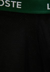 Κοντινή εικόνα μαύρου υφάσματος με υφή και πράσινη ζώνη μέσης που δείχνει μερικό λευκό κείμενο "OSTE" και "L".