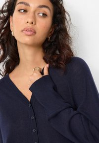 Marineblauer Cardigan mit V-Ausschnitt, Knopfverschluss, gerippten Bündchen und weichem Stoff, getragen über einer Hand, die auf der Brust ruht.