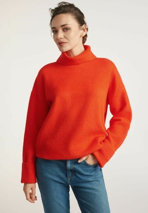 Czerwony, oversize sweter z wysokim, prążkowanym kołnierzem i prążkowanymi mankietami. Miękka tekstura i luźny krój, noszony z niebieskimi dżinsami.