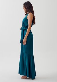 Teal maxi dress made of smooth fabric; sleeveless design with a tied waist and flared hem, featuring a flowy silhouette.