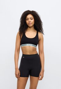 Femme aux cheveux bouclés portant un soutien-gorge de sport noir avec le logo "Lacoste" et un short de cyclisme noir taille haute assorti, sur un fond uni.