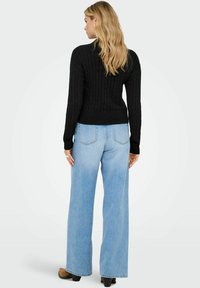 Juodas megztinis su ilgomis rankovėmis, raštuotas kabliukais, derinamas su šviesiai mėlynomis plačiomis kelnėmis. Juodi odiniai batai užbaigia įvaizdį.