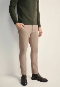 Beżowe spodnie slim-fit w połączeniu z ciemnozielonym swetrem, noszone z brązowymi botkami za kostkę. Gładka tkanina z bocznymi kieszeniami i dopasowanym krojem.