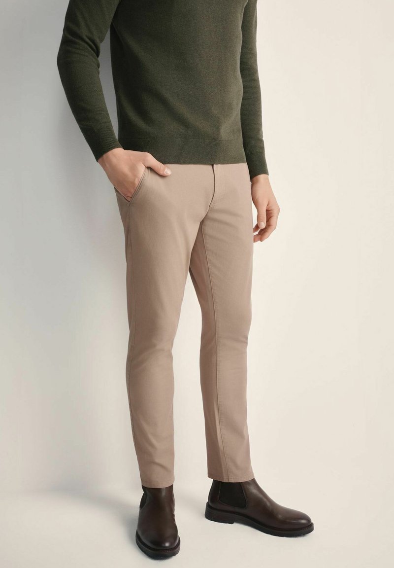 Beżowe spodnie slim-fit w połączeniu z ciemnozielonym swetrem, noszone z brązowymi botkami za kostkę. Gładka tkanina z bocznymi kieszeniami i dopasowanym krojem.
