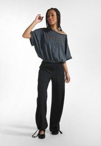 Parte superior suelta de color gris carbón con diseño de hombros descubiertos y logo, combinada con pantalones negros de pierna ancha, con cordones en los bajos y bolsillos.