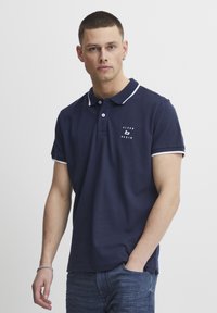 Mornarska polo majica z belim ovratnikom in akcenti na rokavih, z majhnim logotipom na prsih. Narejena iz gladkega materiala, kratke rokave.