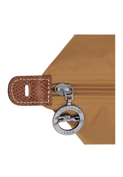 Eck eines braunen Nylonbeutels mit einem strukturierten Lederanhänger, silbernem Reißverschluss und einem runden Metallanhänger, der mit "LONGCHAMP 1948" graviert ist.