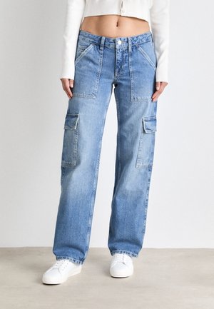 Persoon draagt lichtblauwe wijde cargo-jeans met grote voor- en zijzakken, witte sneakers en een cropped witte trui.