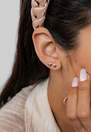 Gold heart-shaped stud earrings on a person's earlobe. Hair is styled with a braided headband. Nails have a sparkly finish.