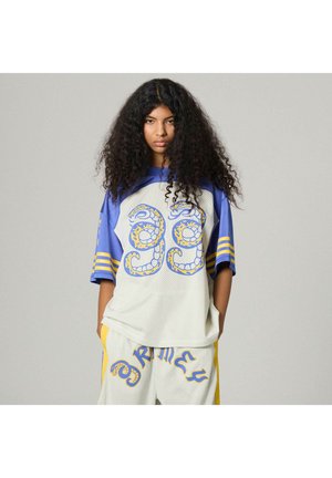 Giovane donna con lunghi capelli ricci che indossa una maglia sportiva bianca e blu con disegni gialli e pantaloncini abbinati, in piedi davanti a uno sfondo grigio uniforme.