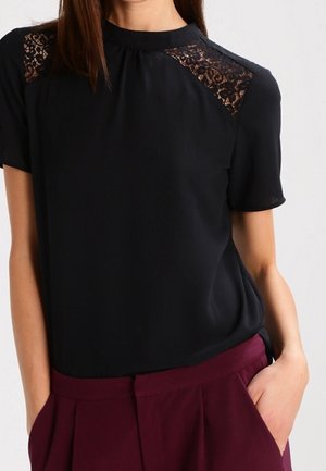 Vrouw draagt een zwart kortermig blouseje met kanten schouderstukken, ingestopt in hoge taille, wijd uitlopende bordeauxrode broek, handen in de zakken.
