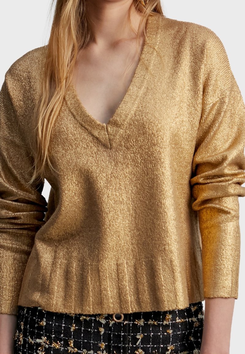 Femme portant un pull col V doré brillant à manches longues et un pantalon noir à motifs avec des accents dorés.