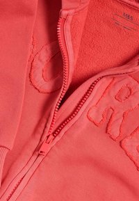 Korálová mikina na zip s měkkou vnitřní podšívkou, texturovaným logem a barevným zipem. Hladká, elastická tkanina s žebrovanými detaily.