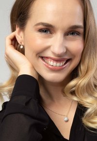 Goldkette mit einem runden Anhänger, der aus einer Gruppe klarer Steine besteht. Das Model hat schulterlange wellige Haare und trägt Ohrstecker.
