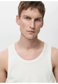 Witte geribbelde tanktop met een ronde halslijn en brede schouderbanden, gedragen door een model tegen een neutrale achtergrond. Zachte textuur zichtbaar.