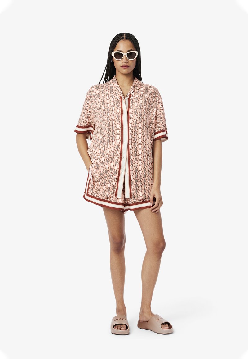 Ensemble de chemise à manches courtes et de short à motifs en crème avec un design géométrique rouge et des finitions. Associé avec des sandales roses claires portant un logo.