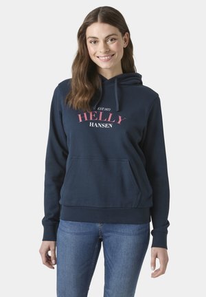 Kobieta z długimi brązowymi włosami, uśmiechająca się, ubrana w granatową bluzę z kapturem Helly Hansen i niebieskie jeansy na jednolitym jasnym tle.