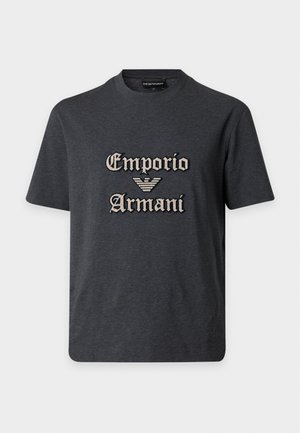 Νέος άνδρας που φοράει μαύρο t-shirt Emporio Armani, σκούρο γκρι τζιν και μαύρα αθλητικά με λευκές σόλες σε λευκό φόντο.