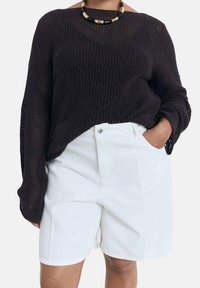 Pull en maille noire avec manches larges et tissage ouvert, associé à un short blanc taille haute. Porte un collier épais noir et or.