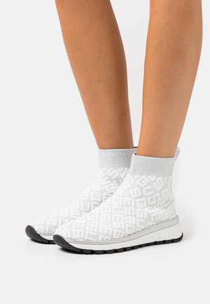 Pieds portant des baskets chaussettes blanches montantes avec un motif de logo texturé, des bracelets argentés à la cheville et des semelles en caoutchouc noir sur fond blanc.