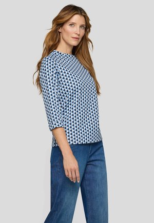 Woman with long red hair wearing a blue and white geometric patterned top and blue jeans, standing against a plain gray background.