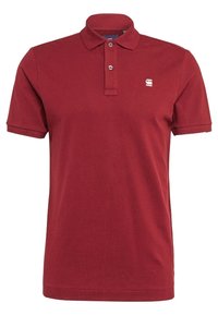 Röd polo tröja i stickad tyg, med krage, tre knappar och en liten logotyp på vänster bröst. Korta ärmar, normal passform.