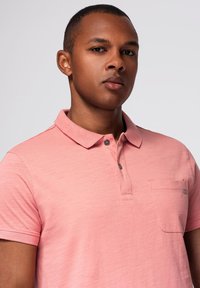 Krátký rukáv, světle růžová polo košile s límcem na knoflíky, jednom kapsičkovým prostorem a jemnou strukturou. Obsahuje detaily loga na kapsičce.