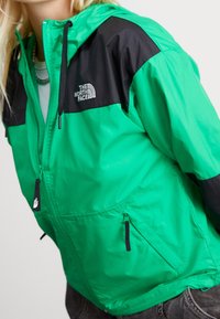 Zelená a černá větruodolná bunda s předním zipem, dvěma postranními kapsami a nastavitelnou kapucí. Na bundě je logo North Face.