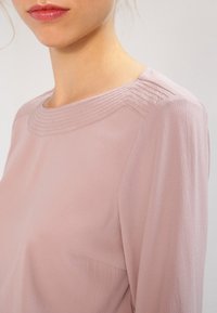 Donna che indossa una blusa rosa chiaro con dettagli cuciti lungo il collo arrotondato e maniche lunghe.