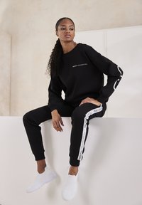 Schwarzer Sweatshirt mit "FRENCH CONNECTION"-Text, kombiniert mit schwarzen Jogginghosen mit weißen Seitenstreifen. Weiße Slip-On-Sneaker. Glattes Material.