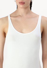 Camiseta de tirantes blanca con escote en el hombro y tirantes finos. La tela es suave y ajustada, con un diseño minimalista sin patrones adicionales.