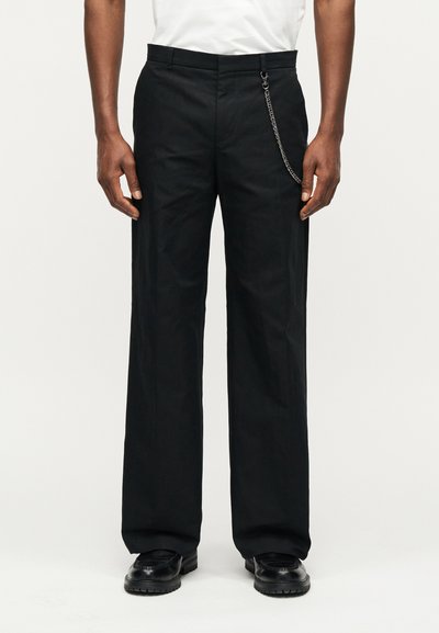 Άνδρας που φοράει μαύρο παντελόνι ίσιας γραμμής με αλυσίδα στο πλευρό, μαύρα παπούτσια και λευκό πουκάμισο, στέκεται μπροστά σε απλό φόντο.