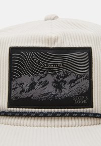 Világos krémszínű kordbársony sapka, fekete folttal, hegyek grafikájával, a "THE DOLOMITES" felirattal és díszes fonott szalaggal.
