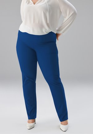 Frau trägt weiße Bluse, eng geschnittene königsblaue Hose und weiße Schuhe mit spitzer Spitze, steht mit den Händen in der Hüfte vor grauem Hintergrund.