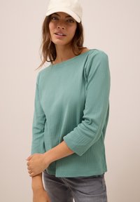 Femme portant une casquette beige clair et un haut à manches longues côtelé teal, posant avec les bras croisés devant un fond beige uni.