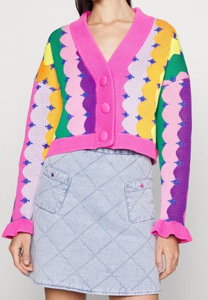 Frau trägt eine gekürzte, mehrfarbige Strickjacke mit Muschelmuster und pinken Knöpfen sowie einen hellblauen, gesteppte Minirock mit Fronttaschen.