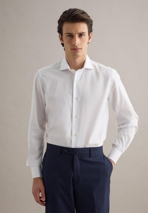 Jeune homme portant une chemise blanche à manches longues rentrée dans un pantalon bleu marine foncé, debout avec une main dans la poche devant un fond uni.